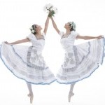 Two dancers wearing pointe shoes hold flowers and reach up, joining hands.