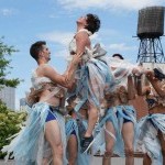During a sunny day, sky bright light blue, a group of dancers wearing plastic bag costumes lift a female dancer on the air.  