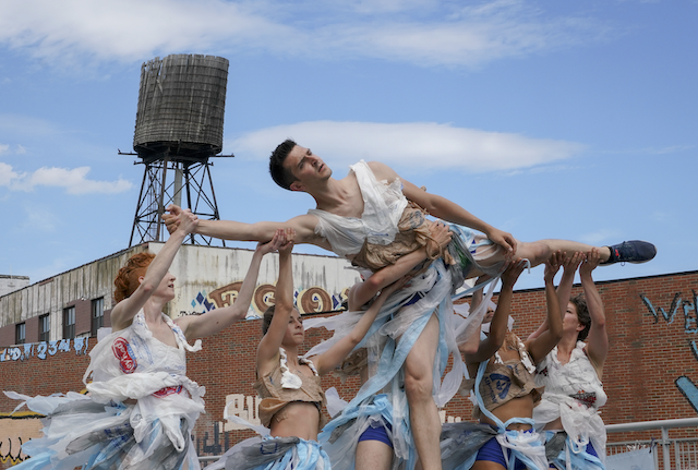 During a sunny day, sky bright light blue, a group of dancers wearing plastic bag costumes lift a female dancer on the air.