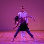 dancer in front in blue and grey wide stance, pulling downward, dancer in white standing quietly behind her.