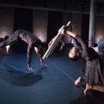 Two dancers, one holding a wooden board and leaning backwards, one in the air.