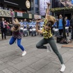 Two dancers side by side with one arm bent and one arm up straight, their head looking down, and one leg straight and one bent