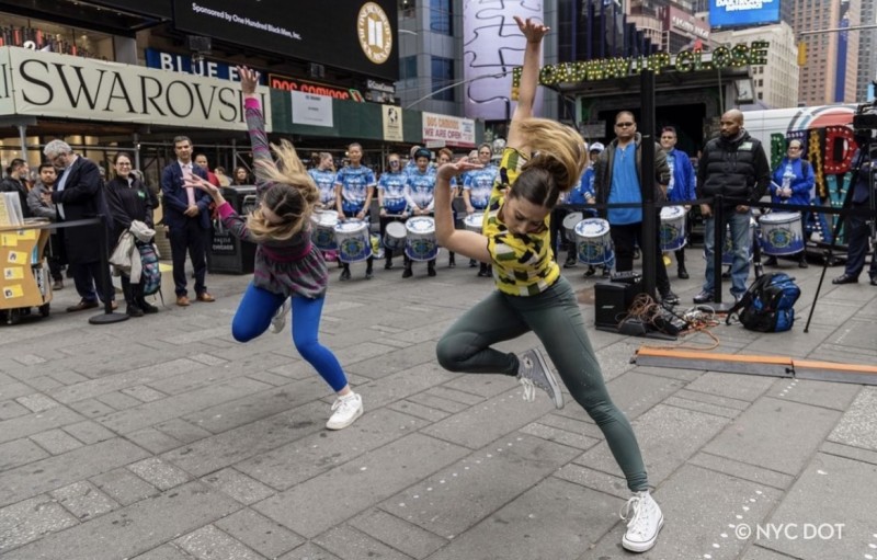 Two dancers side by side with one arm bent and one arm up straight, their head looking down, and one leg straight and one bent