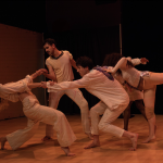Five dancers in neutral costumes perform an interconnected, dynamic movement sequence. 