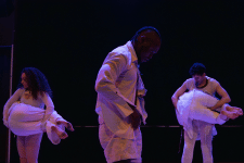 Five dance in white costumes perform on a dimly lit stage, illuminated by blue and purple theatrical lighting.