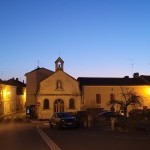 A photo of Mareuil-en-Perigord seen at dusk, with streetlights softly illuminating a church, multiple homes, and parked cars.