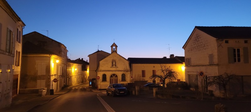 A photo of Mareuil-en-Perigord seen at dusk, with streetlights softly illuminating a church, multiple homes, and parked cars.
