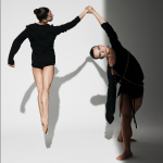 Two dancers holding hands while one jumps in the air facing backwards, wearing a black long sleeve shirt costume.