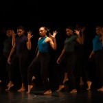 A group of young dancers in dancewear stand in dim light.