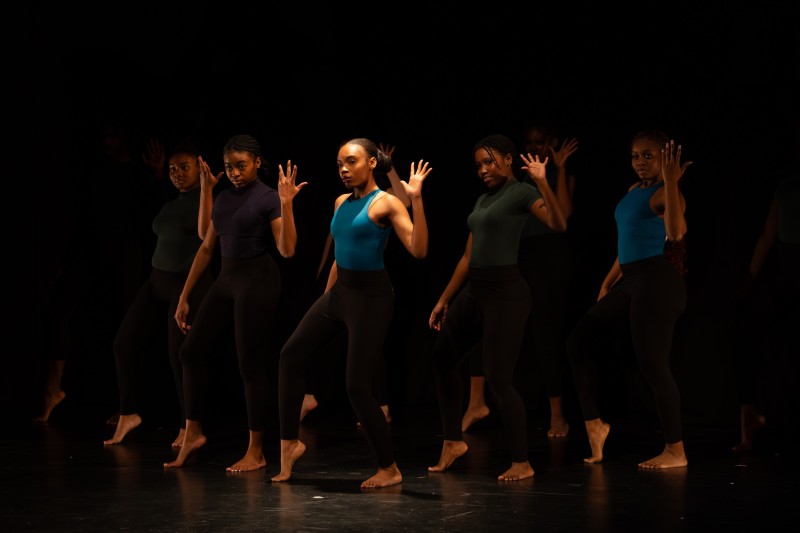 A group of young dancers in dancewear stand in dim light.