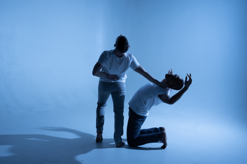 Two men dressed in jeans and t-shirts, one kneels and the other stands with his hand on the other man's neck.
