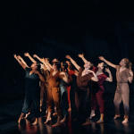 Peridance Collective perform in mixed colors of jumpsuits. Dancers reach one arm to their high right in unison in a dim light.