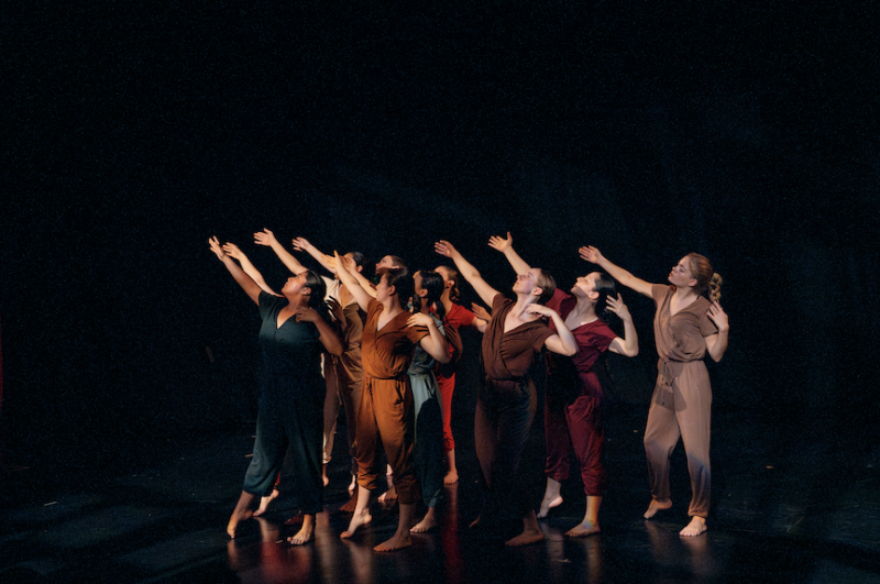 Peridance Collective perform in mixed colors of jumpsuits. Dancers reach one arm to their high right in unison in a dim light.