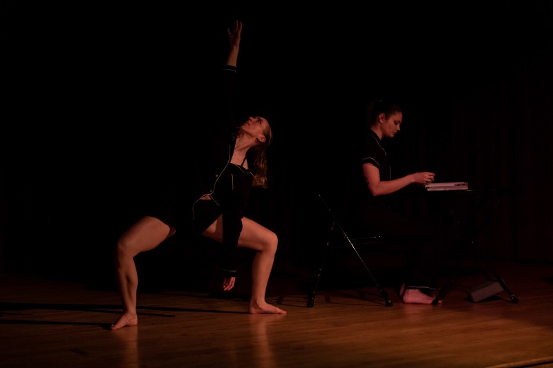 Two dancers - one in a turned out 2nd plie the other sitting at a table with a notebook