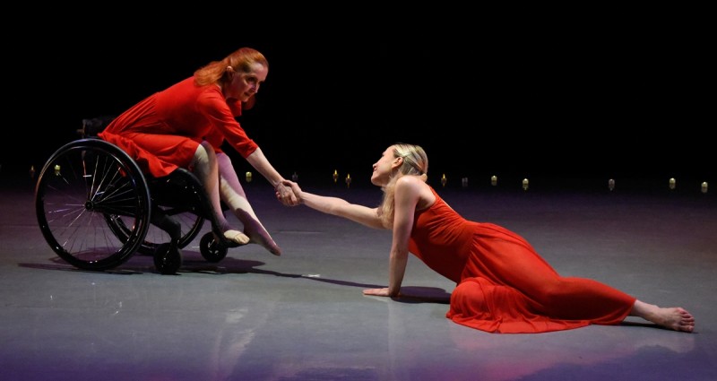 Dancer Kitty Lunn, who uses a wheelchair, and dancer Alison Cook each has one arm outstretched and they grasp hands.