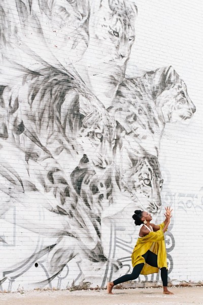 In front of a large tiger mural, a dancer in a yellow flowing robe faces right in a lunge with her hands held in front of her.