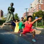 Four Black femmes in colorful dresses hold hands and form a line from a statue of Harriet Tubman pointing into the distance. 