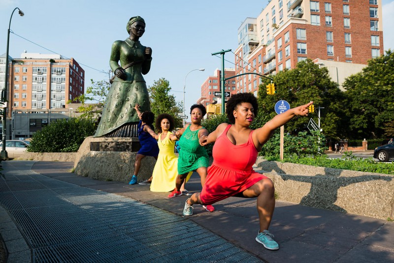Four Black femmes in colorful dresses hold hands and form a line from a statue of Harriet Tubman pointing into the distance. 