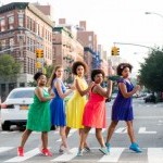 5 people in dresses, green, blue, yellow, pink, purple, stand in the crosswalk and look at the camera with NYC behind them.