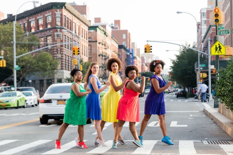 5 people in dresses, green, blue, yellow, pink, purple, stand in the crosswalk and look at the camera with NYC behind them.