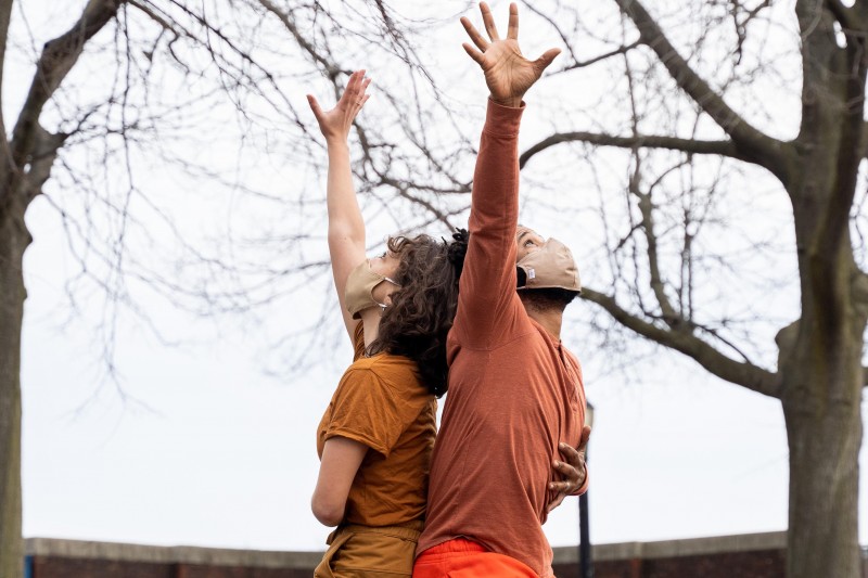 Two dancers reaching towards the sky. Both are wearing masks and wearing muted, neutral colors