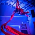 woman in unitard is suspened in the air wrapped in red silk while swirling the silk below
