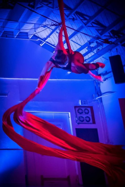 Female Aerial Dancer in unitard is suspended in air and wrapped in red silk the silk is swirling below like a ribbon.
