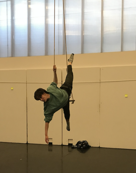 A dancer in casual workout clothing is laying out into space while lying across the bar of a trapeze.