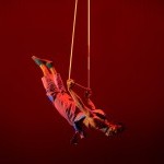 Black Aerial dancer in jumpsuit holds himself horizontally aloft 
