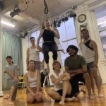 Six smiling people gathered around 1 person sitting in low trapeze, all dressed in dance clothing