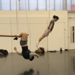 2 people in workout gear on low trapeze; 1 hangs from knees, the other pushes body up on bar with hands in diagonal shape.