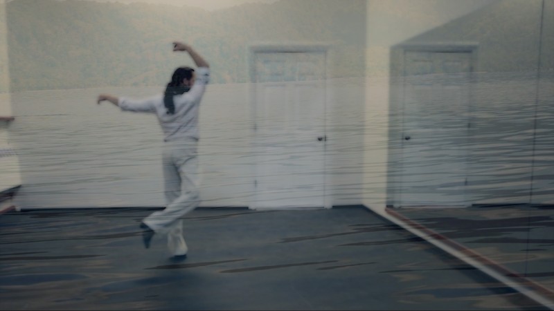 A man holds his arm up, his leg kicked back, as he looks ate a door onto which is projected the image of a calm bay.