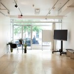 Interior of a studio with white walls and a wood floor. Beyond the glass windows is a car and a passerby looking in.