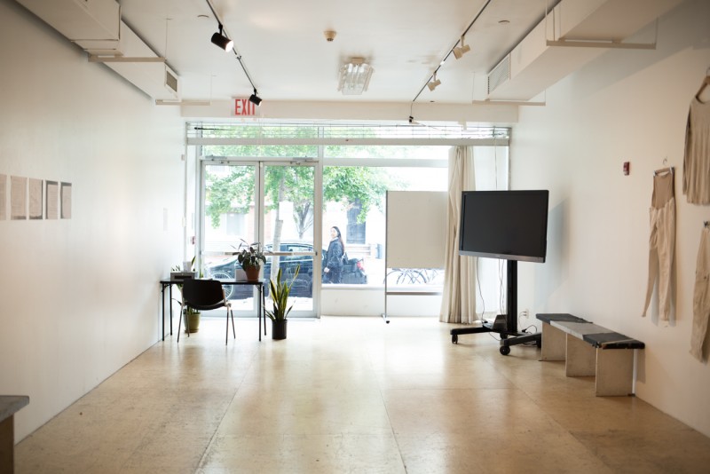 Interior of a studio with white walls and a wood floor. Beyond the glass windows is a car and a passerby looking in.