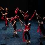 Group of 5 dancers holding bamboo sticks with red ribbon tassels looking up to the left