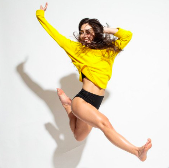 Briana jumping with one leg straight and the other bent. She is in black shorts and a yellow sweater.