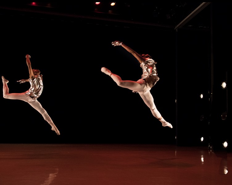 Two male dancers in white and silver costumes in a grand jete leap.