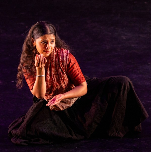 A woman with long dark hair wearing a red-orange tunic and a dark skirt, seated with right hand to chin and left arm crossed.
