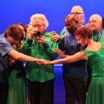 Dancers and musicians costumed in blues and greens embrace each other around a central figure playing a pocket trumpet
