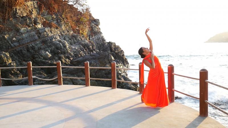 Dancer Yuritzi Govea in a flowing orange dress, by the ocean in Mexico, with her arm reaching to the waves and clouds