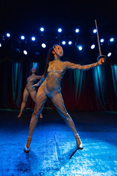 A dancer, wearing a rhinestone bodysuit and rhinestone face cage, holds a sword in a gesture that resembles fencing.