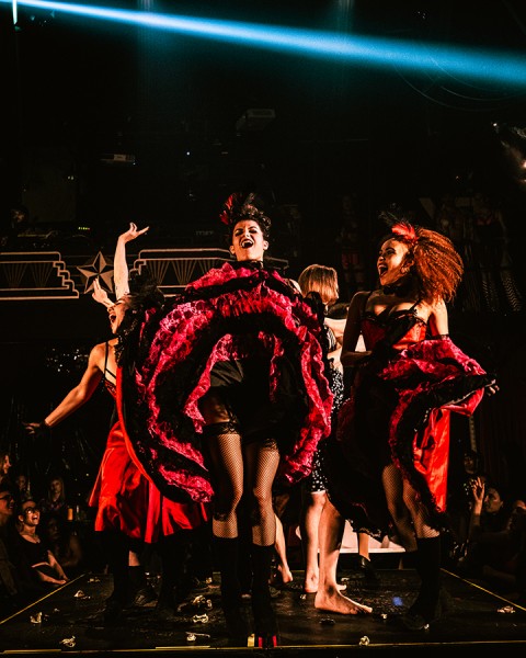 A group of dancers in red /black costumes dance on a stage in the round.