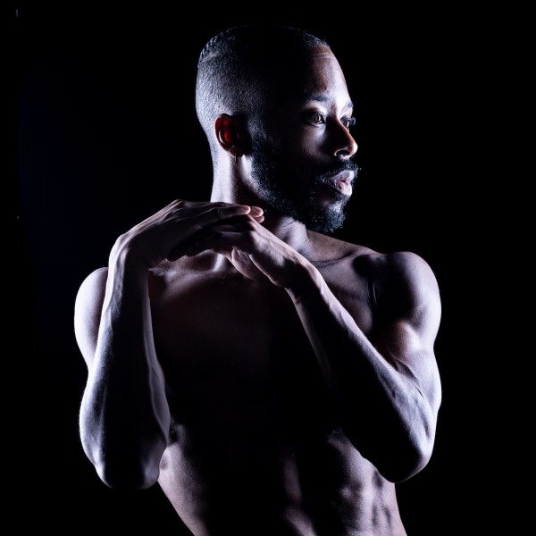 Image of Christian in front of a black backdrop, looking to the left with their hands clasped close to their right shoulder.