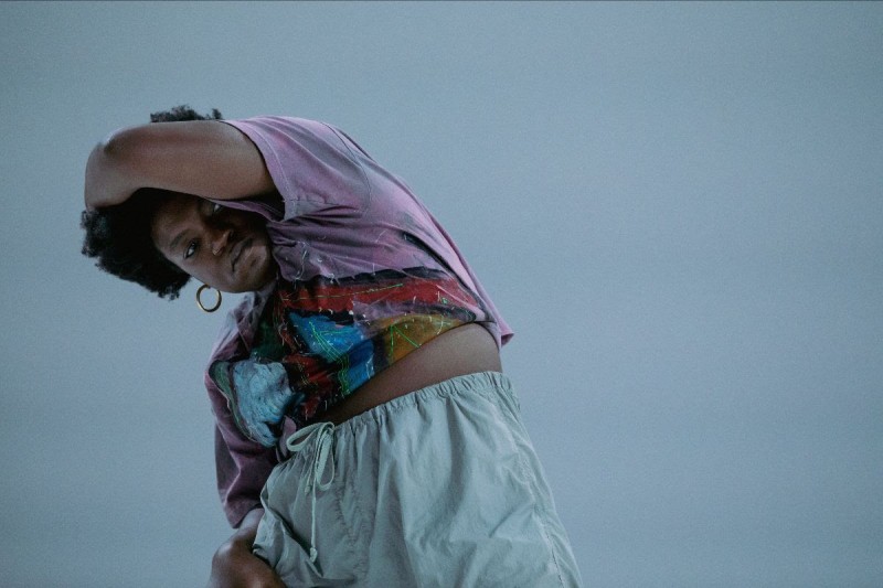 A dark skinned Black woman with a 4c puff of hair looks through her arm into the camera and wearing a purple graffitied shirt.
