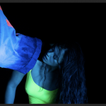 Image of kimiko tanabe in a neon green tank top offering her shoulder to another dancer’s ankle in front of a black background. 