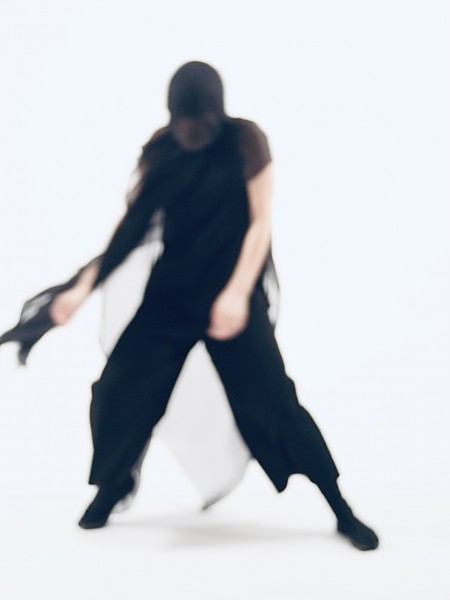 Blurry photo of Yoko Murakami caught mid movement against a white background. Draping black fabric covers her face and body.