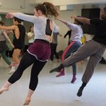 An image of two dancers mid-air in focus, with other dancers in the background of a studio.