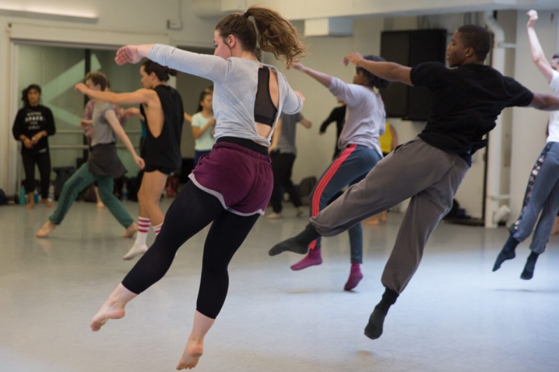 An image of two dancers mid-air in focus, with other dancers in the background of a studio.
