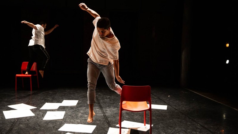 Two men jumping between chairs with paper on the floor.