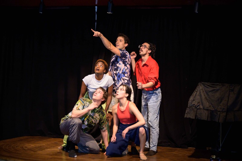 Five performers dressed in vibrant street clothing gape at something off screen, looking amazed. 
